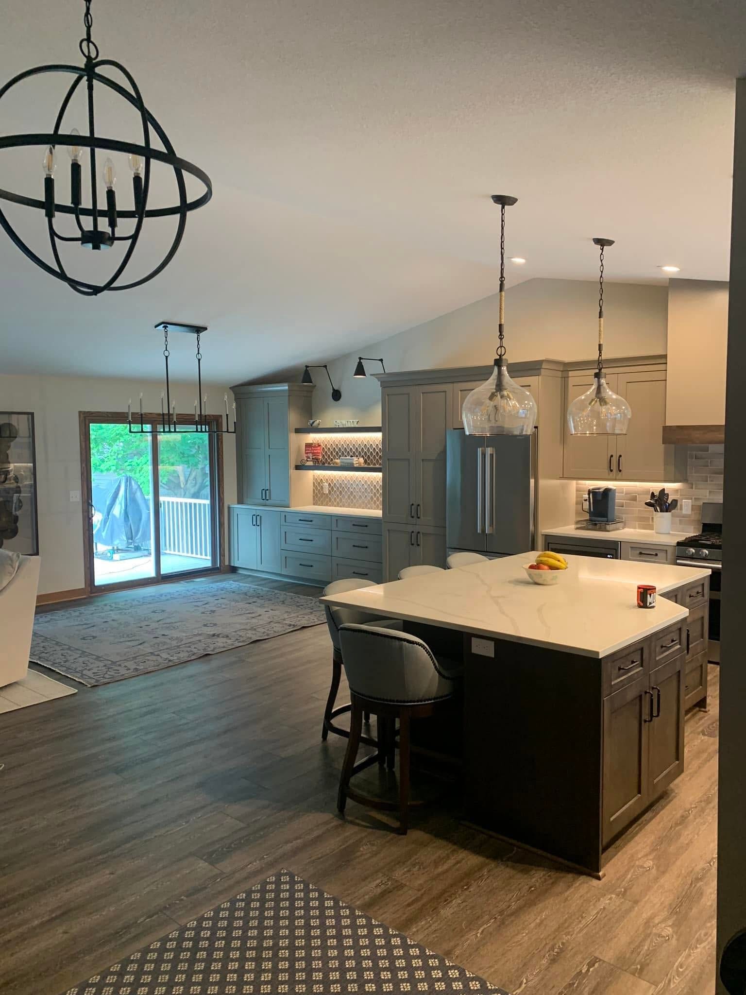 Finished kitchen with dark shaker island, white countertop, glass pendant lights, orb chandelier, light gray cabinetry to the ceiling, stainless refrigerator, and wood-look plank floor