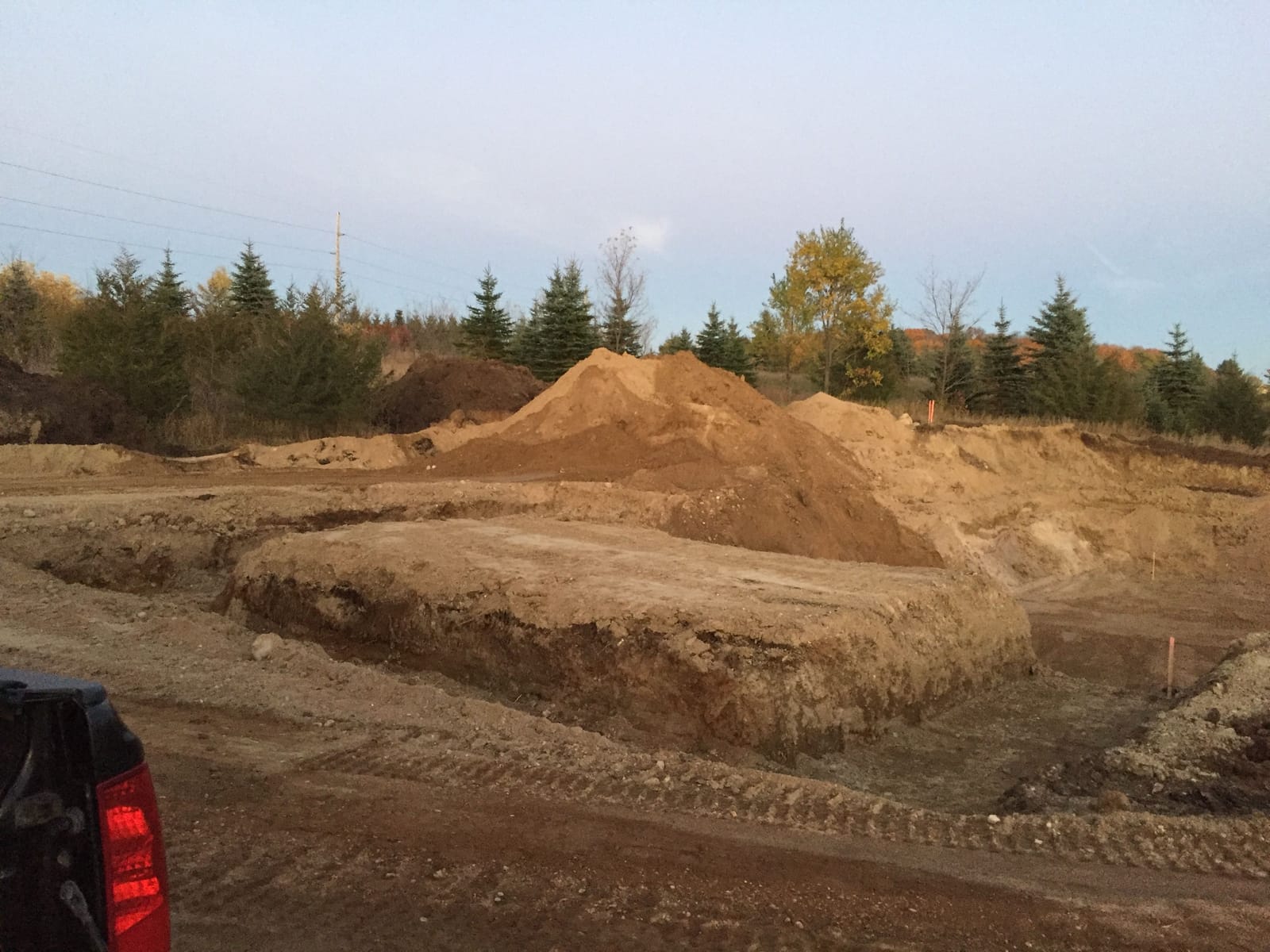 Excavated building site with a dug foundation footprint and stockpiled soil, tree line in the background