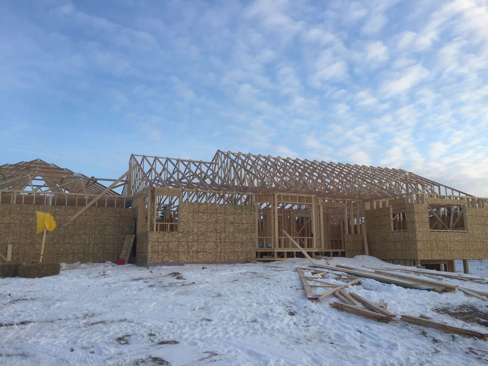 Framed house with roof trusses set across the full width, sheathed walls with window openings, and lumber staged on a snow-dusted site under a blue sky