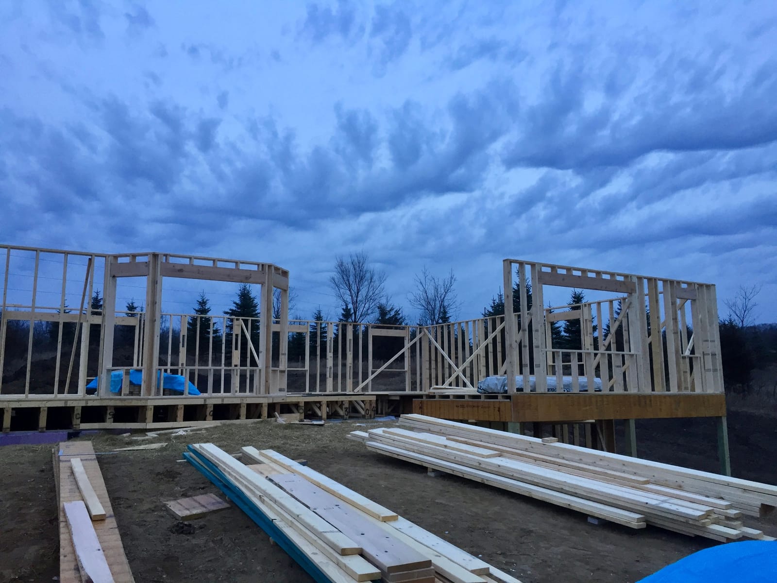 House under construction with exterior walls framed and sheathed, window openings cut, stacked lumber and a blue tarp in the foreground, snow on the ground under a cloudy sky