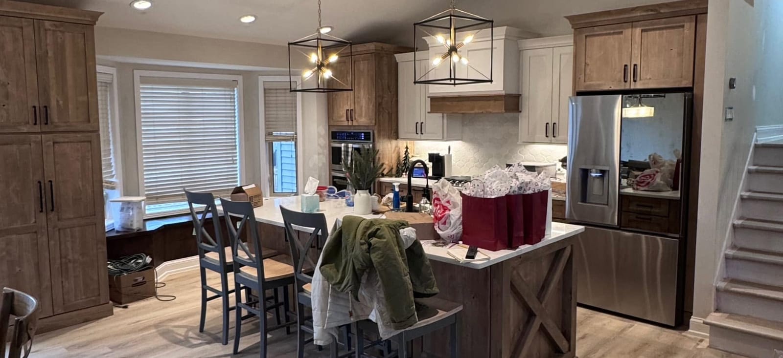 Kitchen after remodel: knotty-alder tall cabinets, white shaker uppers, wood-clad island with X-brace end and white top, cage pendant lights, stainless refrigerator, and wood-look plank flooring