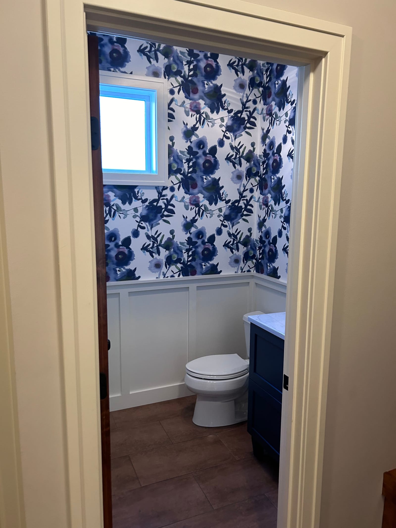 Powder room seen through its doorway: white board-and-batten wainscot with a cap rail, blue floral wallpaper above it, a small window with white trim, a dark vanity and a toilet, on wood-look flooring