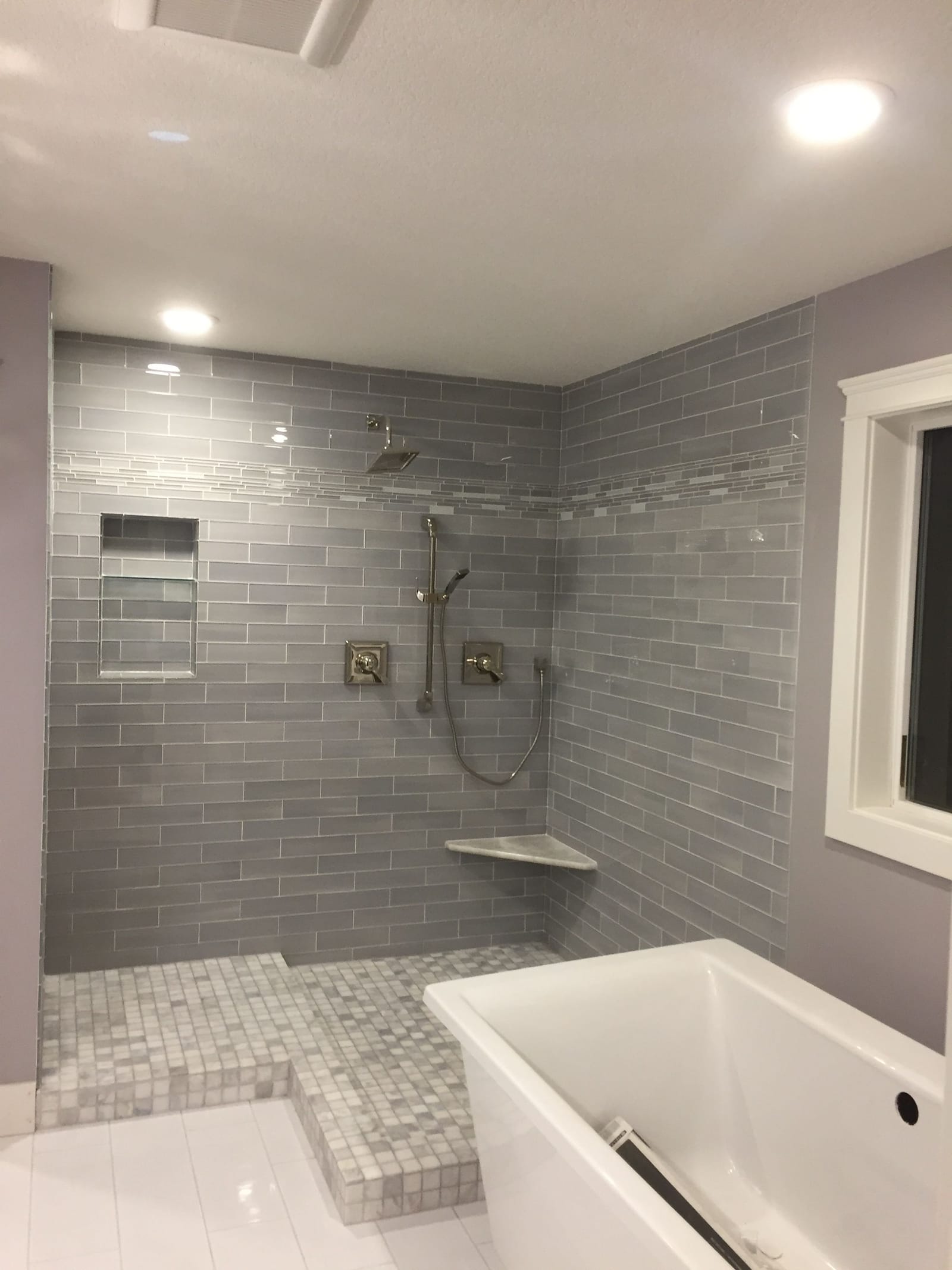 Bathroom with gray subway-tile shower, tiled bench and recessed niche, mosaic tile floor, freestanding white tub beneath a window, and recessed lighting