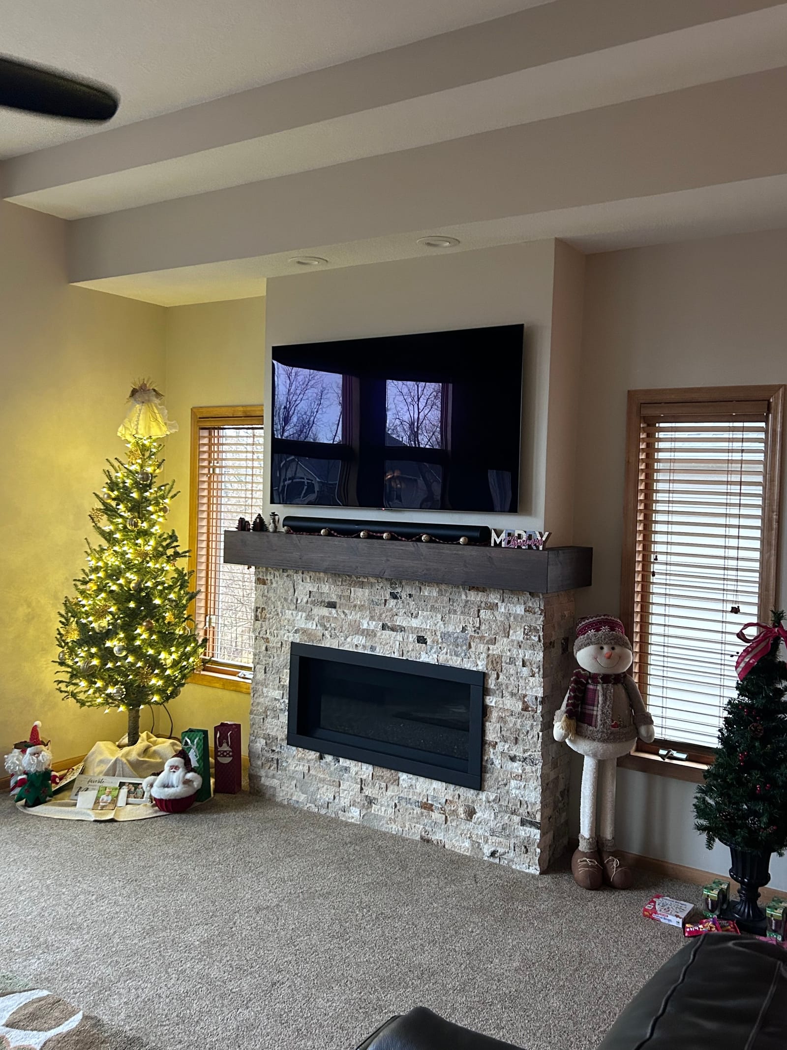 Living room with a floor-to-ceiling stacked-stone fireplace, a linear fireplace insert, a dark wood mantel and a television above it, flanked by windows, decorated for the holidays with a tree and a snowman figure