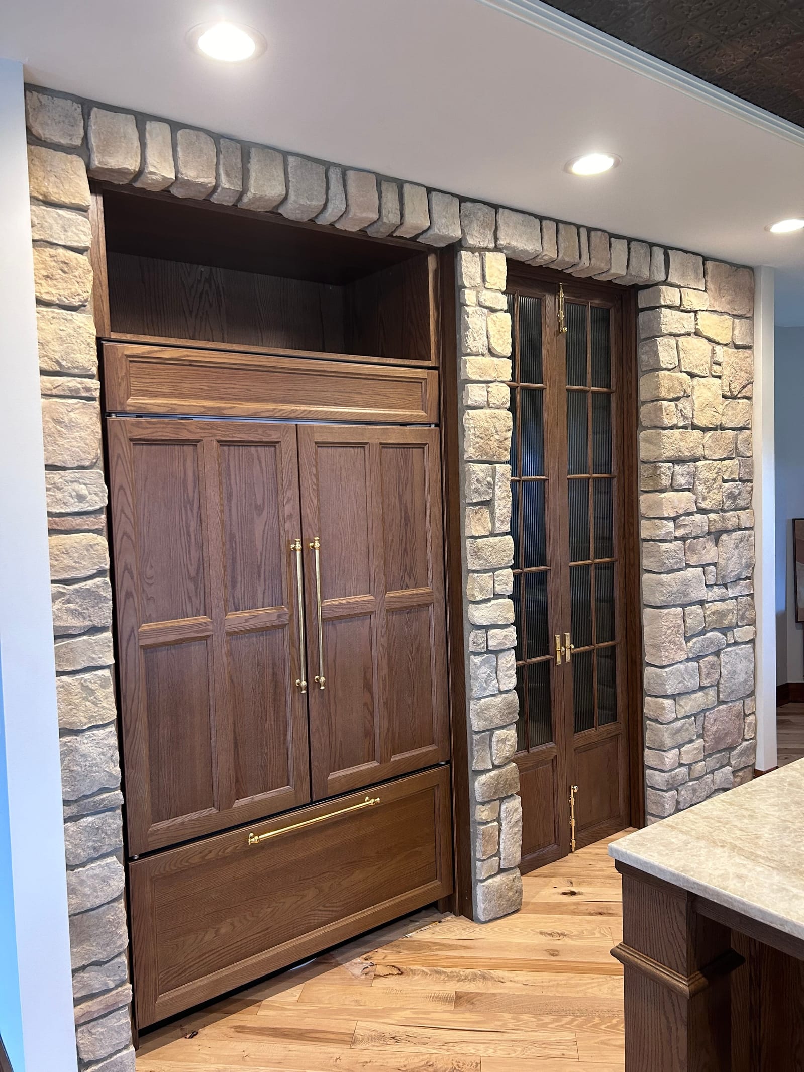 Close view of a stone-veneer kitchen wall with a built-in wood refrigerator panel, wood cabinet doors and a glass-front pantry door, beside a stone-topped island