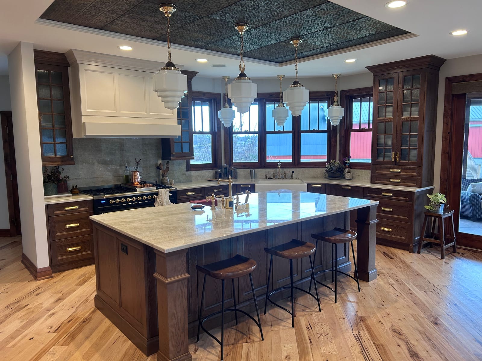Kitchen with a large dark-wood island with a light stone top and four stools, a white range hood over a black range, glass pendant lights, a pressed-tin coffered ceiling panel, dark cabinets and a hardwood floor