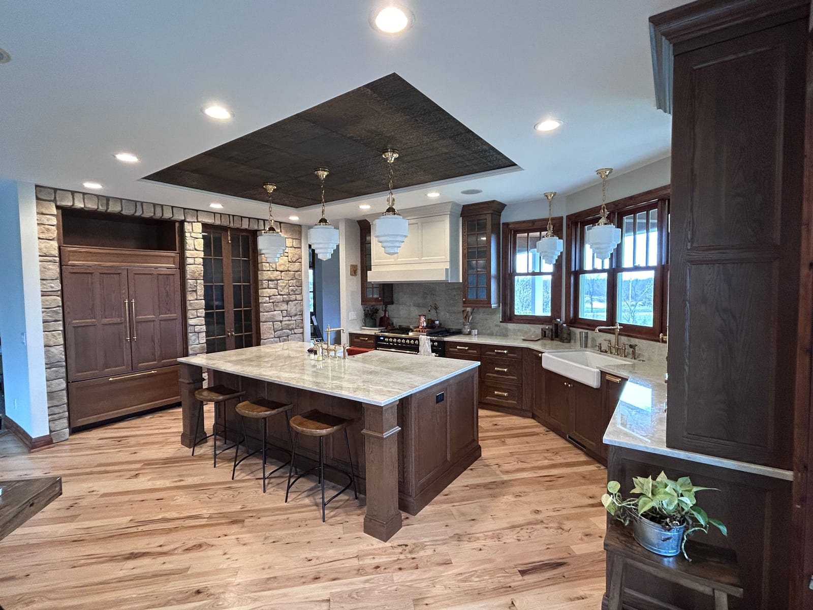 Wide view of a kitchen with a stone-veneer wall and wood-panelled refrigerator on the left, a dark-wood island with a stone top and stools, a white range hood, pendant lights under a pressed-tin ceiling panel, and a hardwood floor