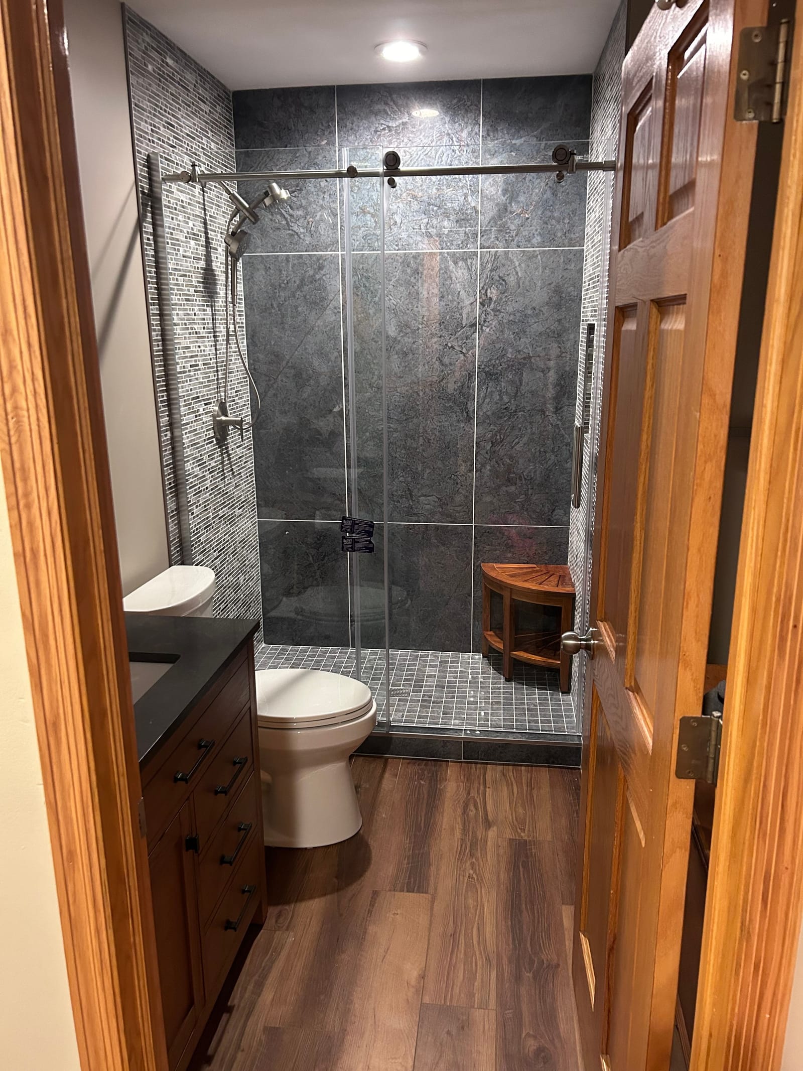 Bathroom with a walk-in shower tiled in large grey-blue tile with a corner seat and a frameless sliding glass door, a dark vanity with a black countertop, and wood-look plank flooring, seen past a stained wood door