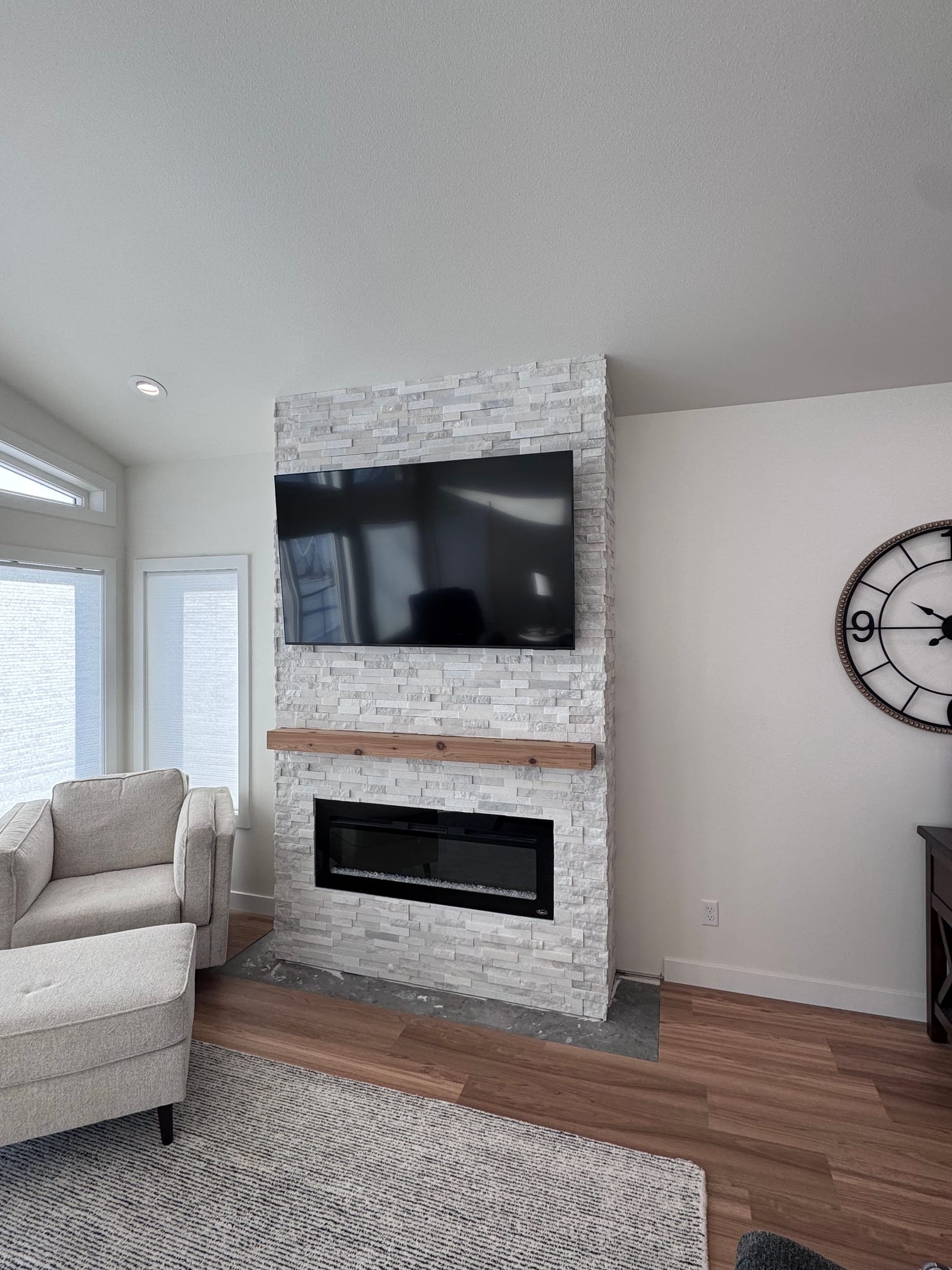 Living room with a white stacked-stone fireplace wall, a linear fireplace insert, a natural wood mantel, a television above, a round wall clock, an armchair and wood-look flooring