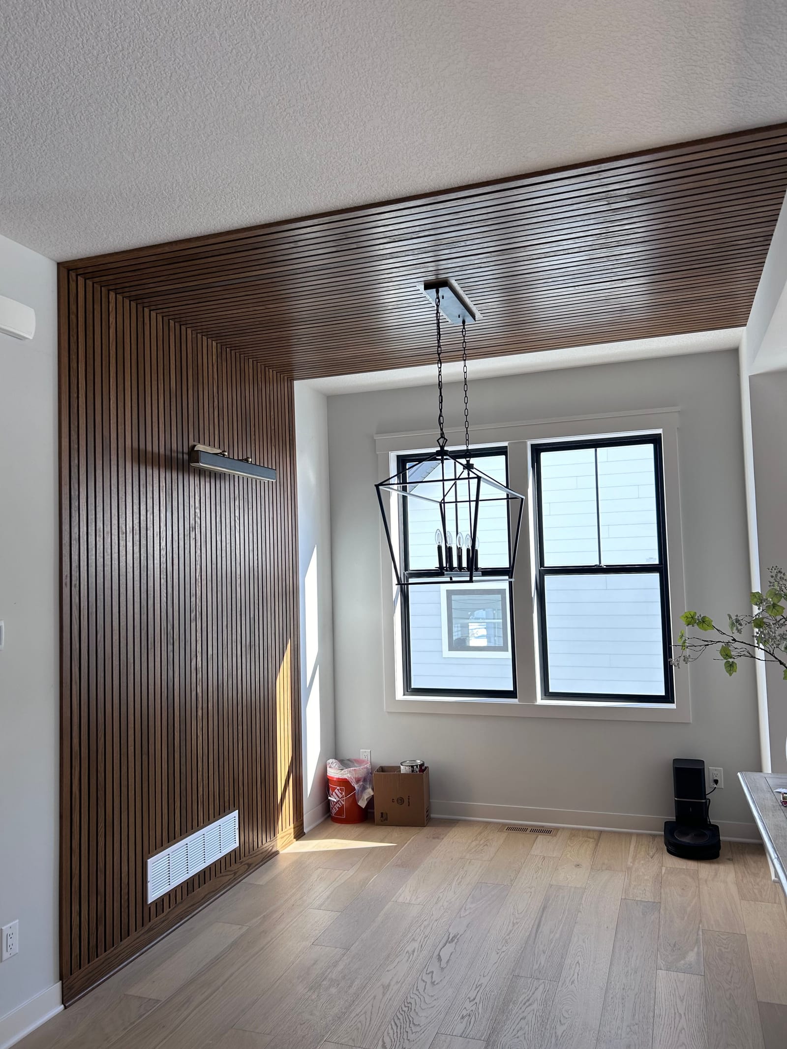 Room corner finished in vertical stained-wood slats that continue across the ceiling of a window bay, black-framed double windows, a lantern pendant and a light hardwood floor